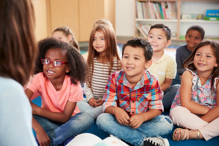 children sit cross-legged and listen attentively to a teacher